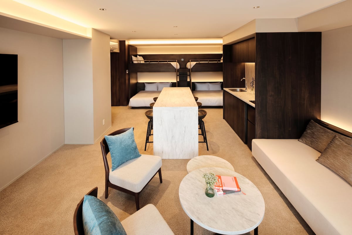 Una habitación de hotel moderna con un sofá, dos sillones, una mesa redonda pequeña, una litera con escritorio y una zona de cocina.