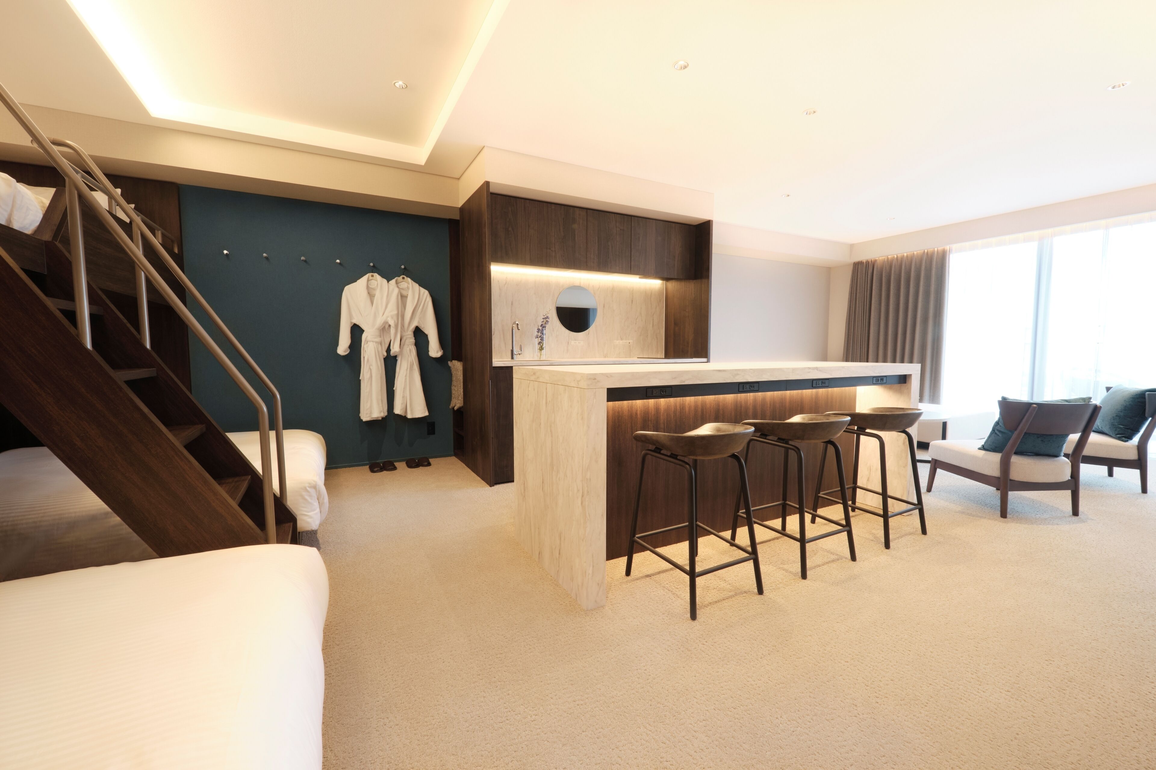 Una habitación de hotel moderna con una escalera, una sala de estar con sofá y sillones, una pequeña zona de bar con taburetes y un amplio ventanal que proporciona luz natural.