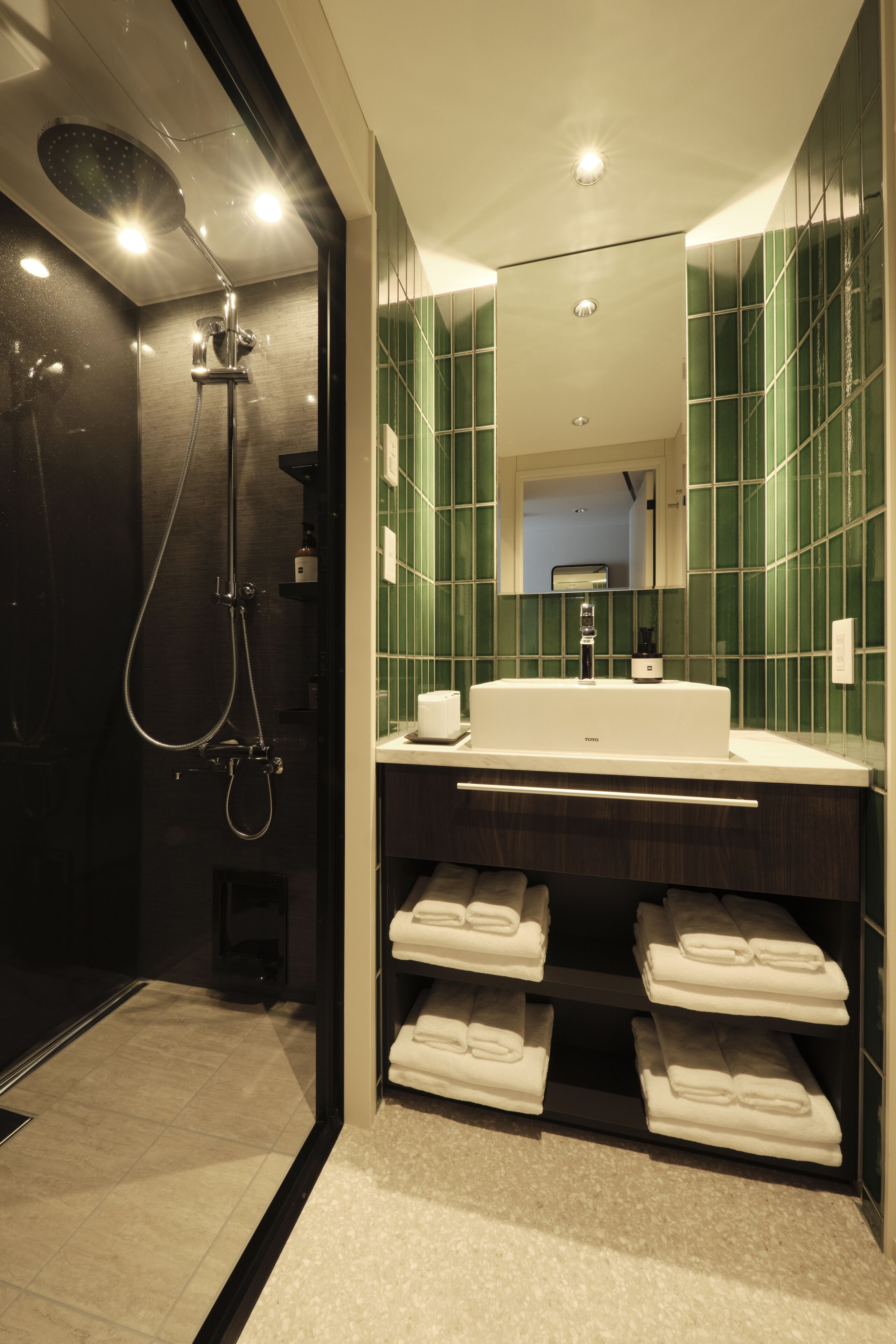 A modern bathroom with a large mirror, green tiled walls, a white sink, and a shower.