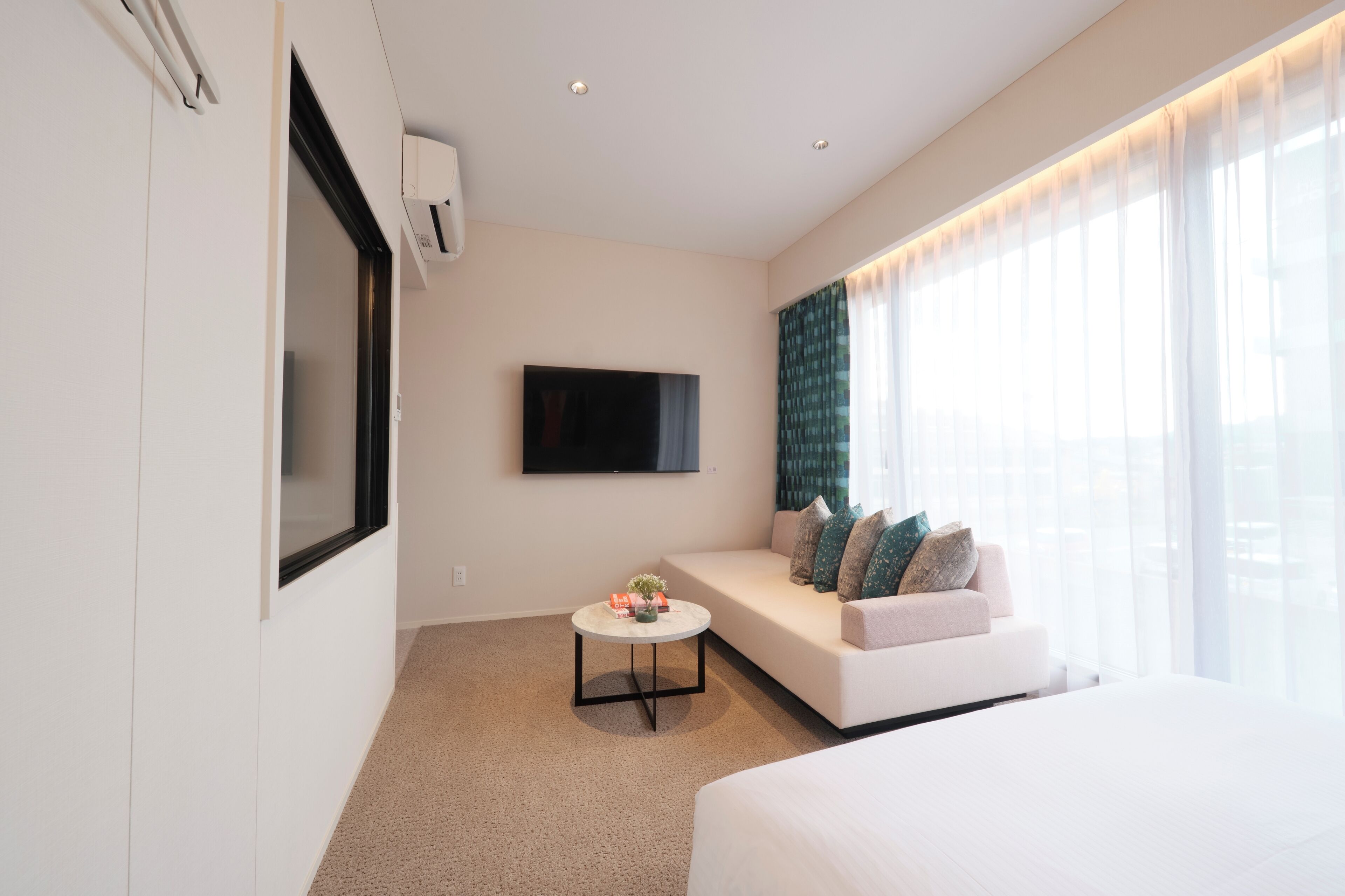 Una habitación de hotel moderna con una cama grande, un sofá, una pequeña mesa de centro, un televisor de pantalla plana montado en la pared y un amplio ventanal con cortinas translúcidas.
