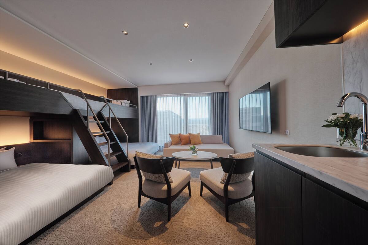 Habitación de hotel moderna con literas, un sofá, dos sillones, una mesa de centro, una zona de cocina con lavamanos y un ventanal con cortinas.