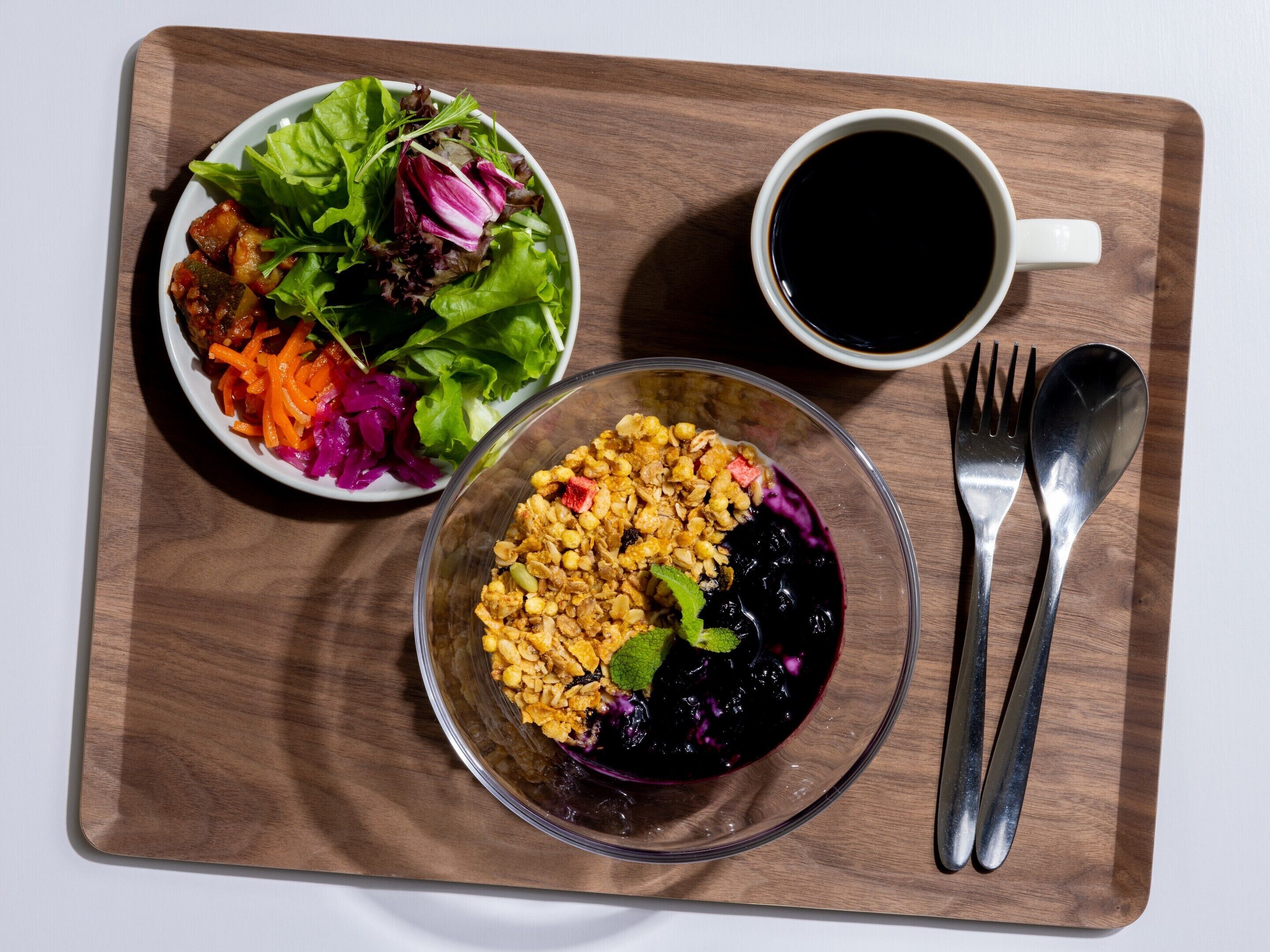 Un repas composé d’une salade, d’un bol de granola aux fruits rouges et d’une tasse de café noir, le tout servi sur un plateau en bois.