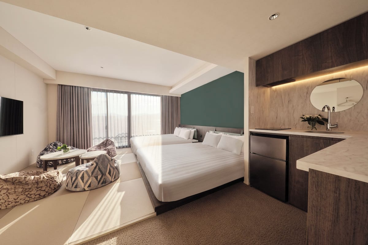 Habitación de hotel moderna con una cama grande, un escritorio, una kitchenette pequeña y un ventanal con cortinas.