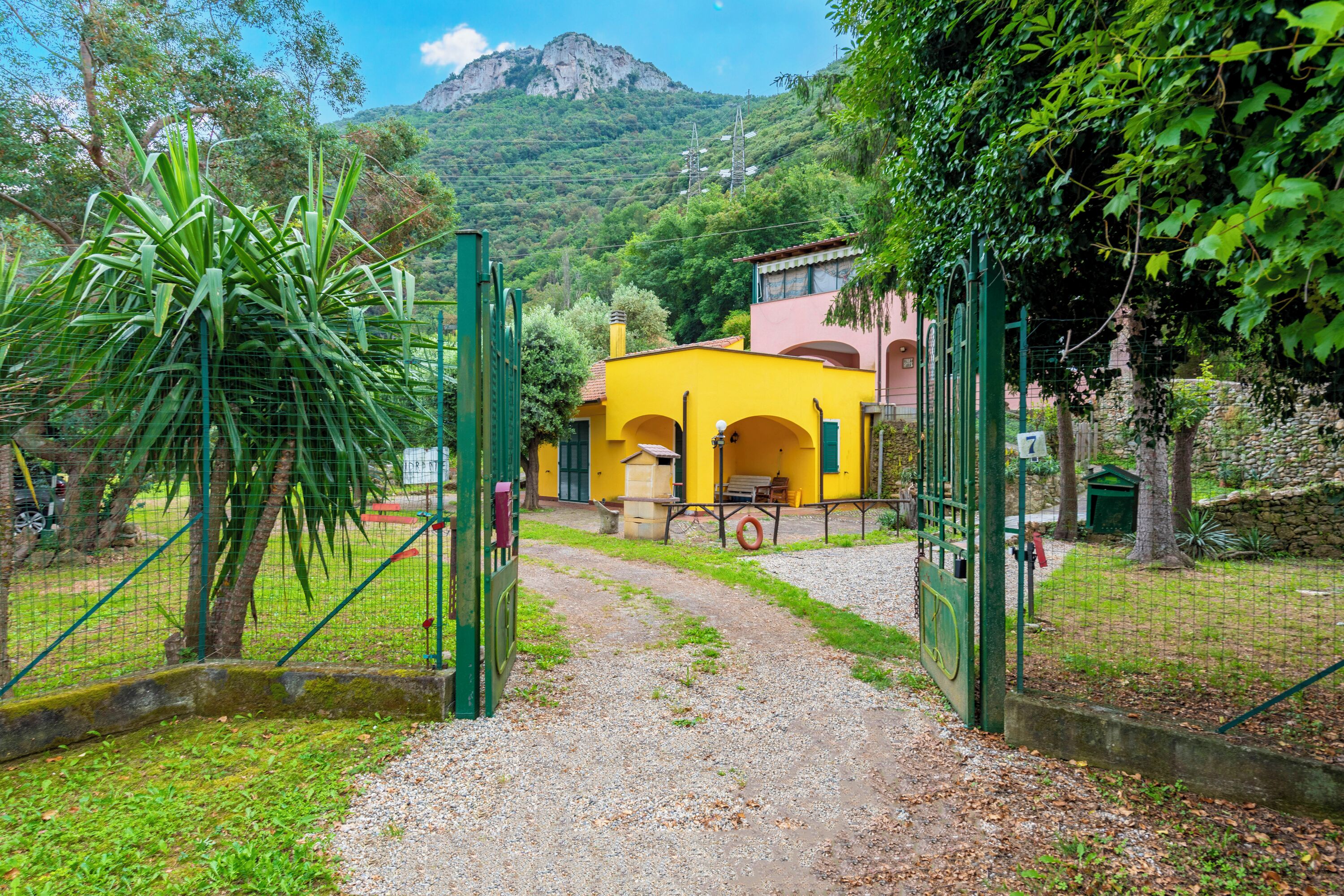 Un chemin mène à un bâtiment jaune orné d’arcades, entouré de verdure et avec une montagne en arrière-plan.