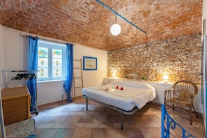 A bedroom with a brick ceiling, a bed, a wicker chair, a window with blue curtains, and a stone wall. - Villa Sara - 5 Min From The Sea (Finale Ligure)