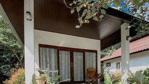 A covered outdoor seating area with a wooden bench and a table, surrounded by large green plants. - Maliblue resort khanom (Khanom)