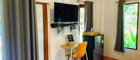 Una habitación compacta con piso de madera, una mesa pequeña con una silla, un televisor montado en la pared y un refrigerador.