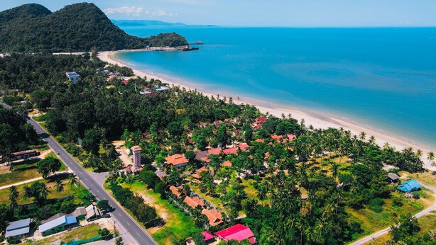 An aerial view of a coastal area with a beach, a road, and residential buildings surrounded by greenery. - Maliblue resort khanom (Khanom)
