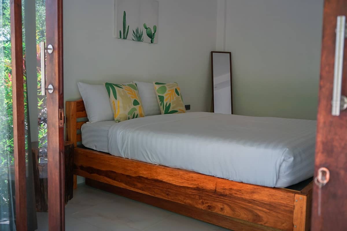 Un lit en bois avec une literie blanche et des coussins à motifs, un tableau encadré représentant des cactus accroché au mur, et un miroir.