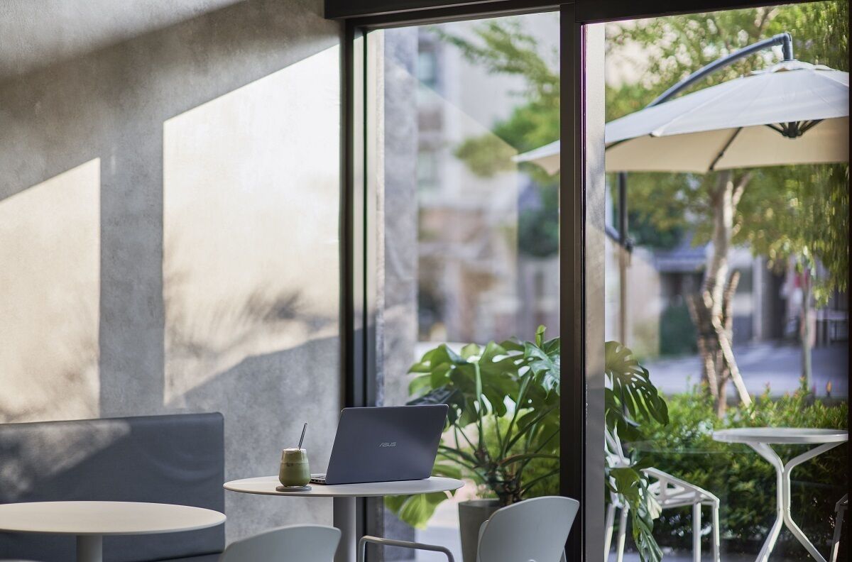 A modern cafe with a laptop on a table, large windows, and outdoor seating with umbrellas.