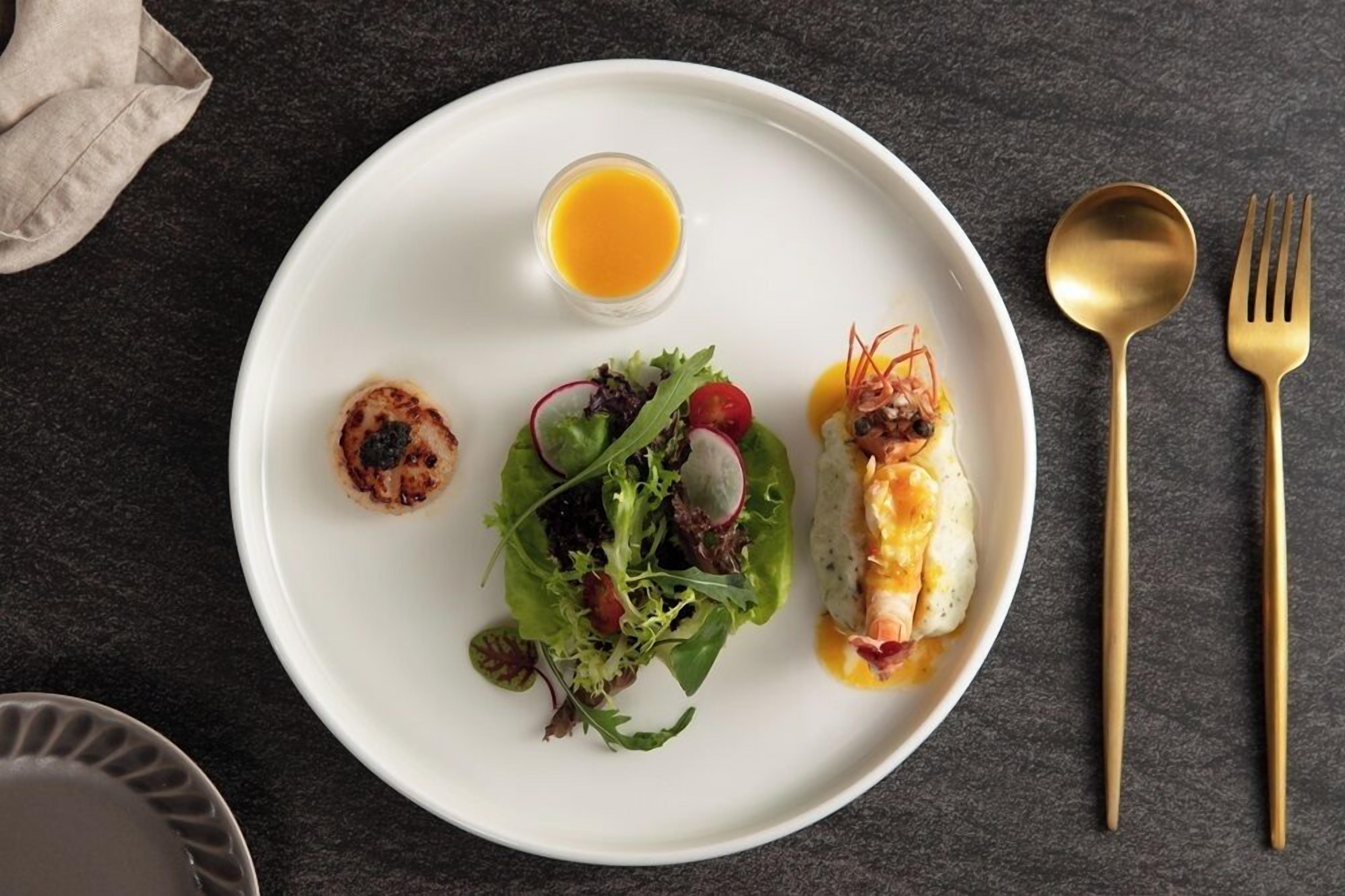 A white plate with a salad, a small dish of sauce, and a glass of orange juice on a dark surface.