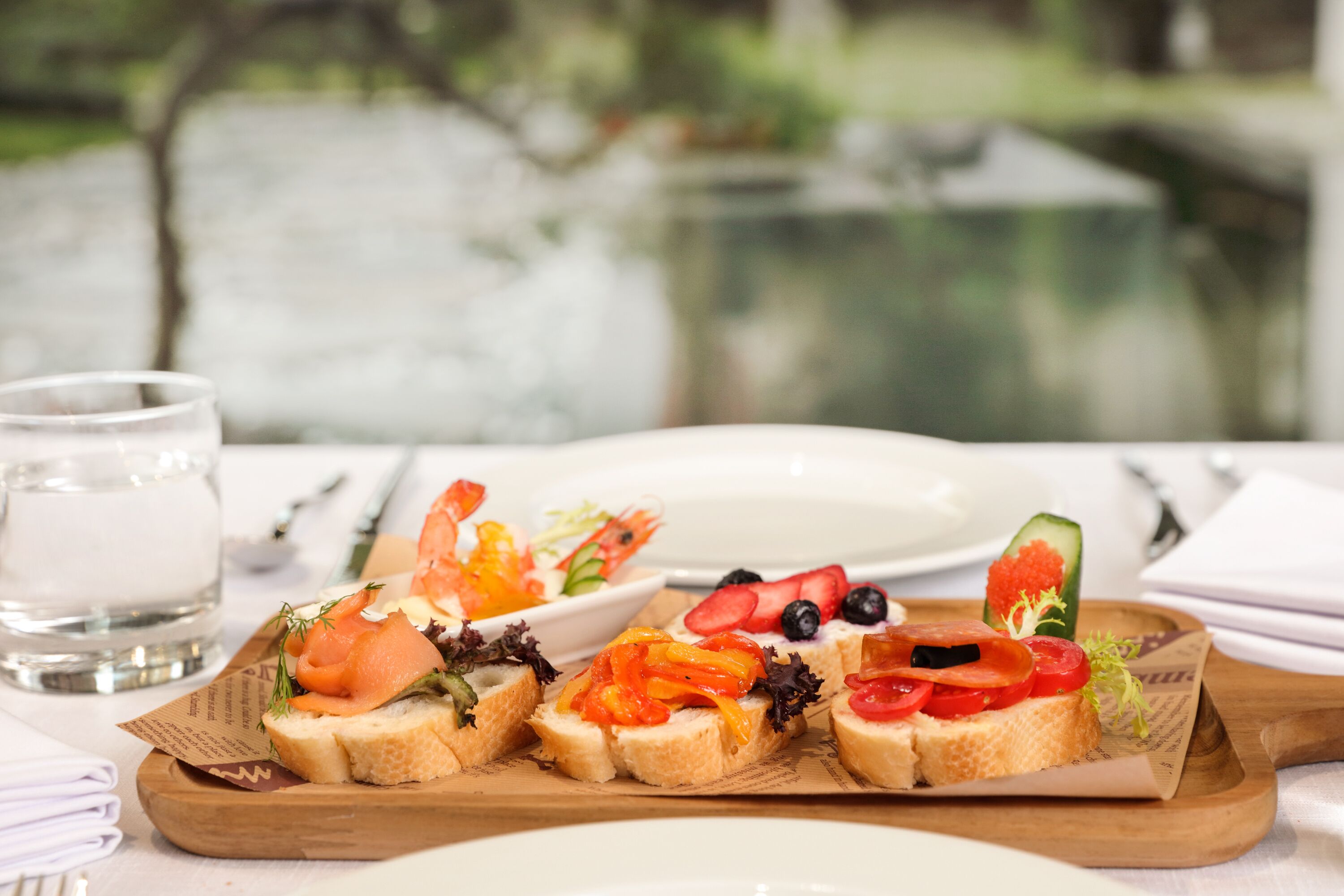 A wooden board with various food items, a glass of water, and a white plate on a table with a blurred background.