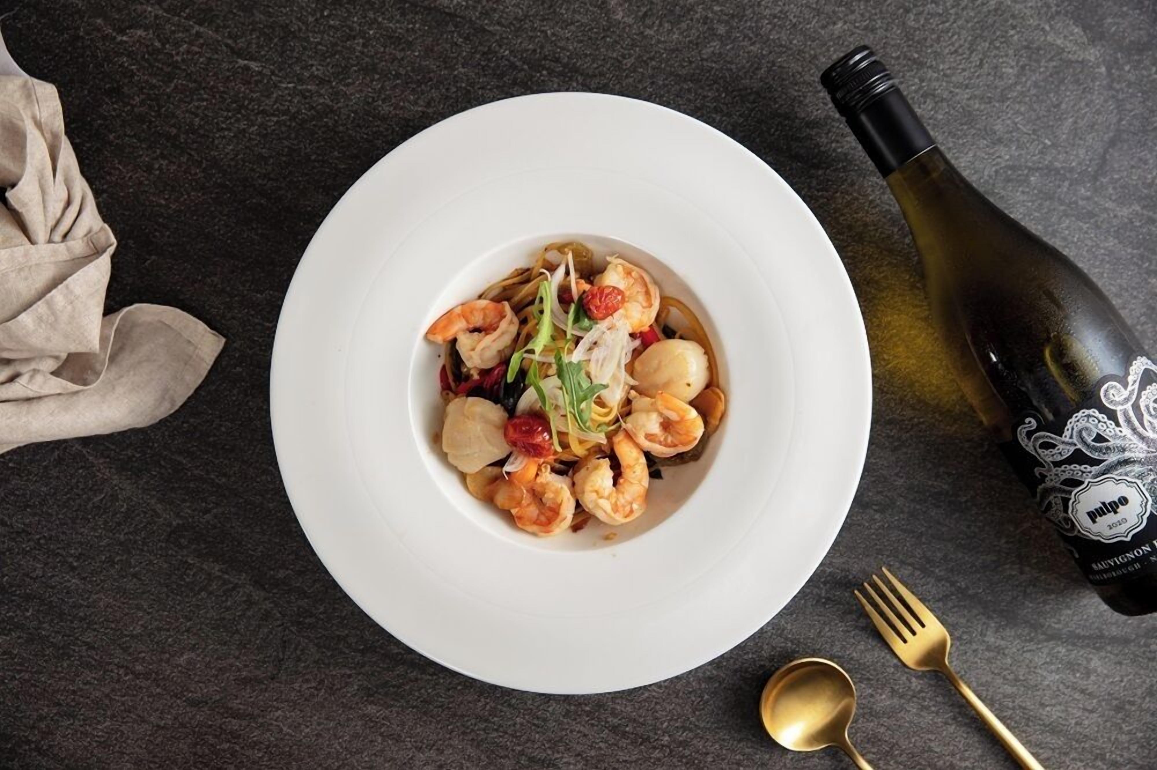 A white plate with a seafood dish, a bottle of wine, and a gold fork and spoon on a dark surface.