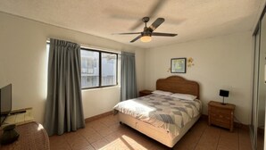 Un dormitorio con cama, ventilador de techo, ventana con cortinas, mesita de noche y un cuadro colgado en la pared.