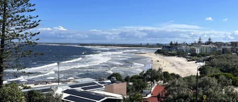 A coastal view with a beach, ocean waves, and buildings in the background.