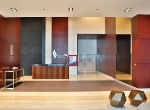 A modern reception area with a wooden reception desk, a glass elevator, and a wall with a logo. - Homesgetaway- 2BR in Marina Gate Tower 2 (Dubai)