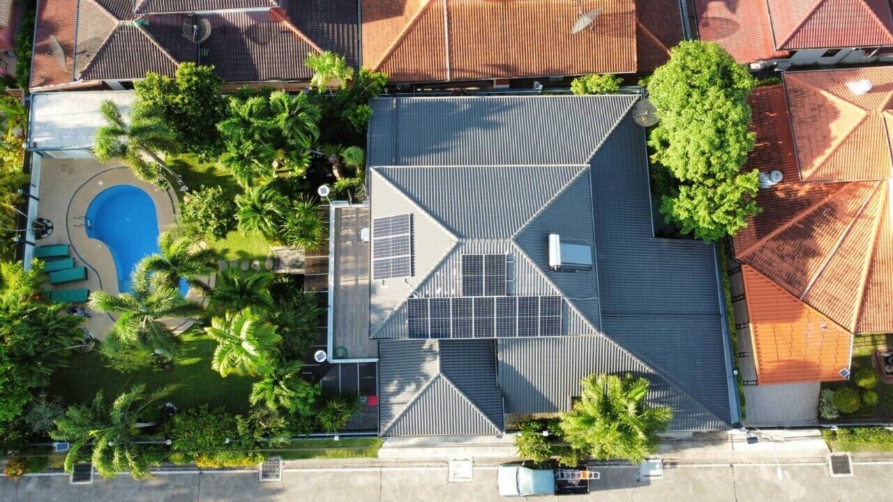 An aerial view of a residential area with a swimming pool, green lawns, and a house with a sloped roof and solar panels.