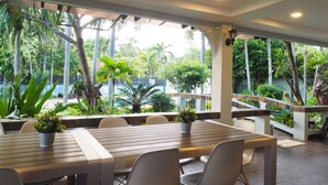 Un área de comedor al aire libre con techo, que incluye una mesa y sillas de madera, rodeada de plantas en macetas y vistas a una piscina y follaje.