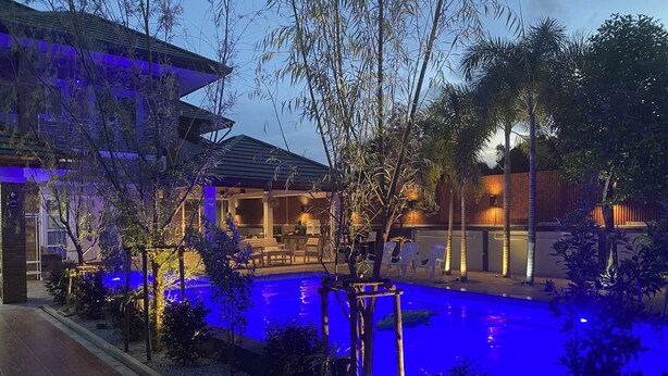 Un área de piscina con una silla de descanso y una mesa de comedor, rodeada de plantas y un edificio con techo inclinado.