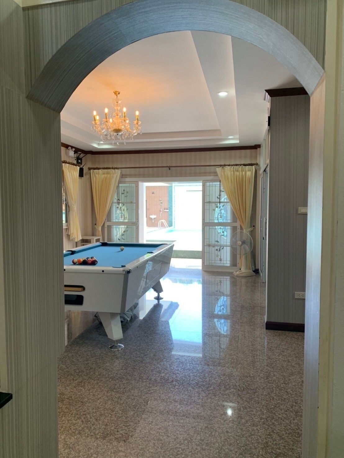 A spacious room with a pool table, a chandelier, and a view of the outside through large windows.