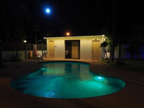 Une piscine à bord incurvé, entourée d’une terrasse avec un petit bâtiment doté de deux portes et d’un banc.