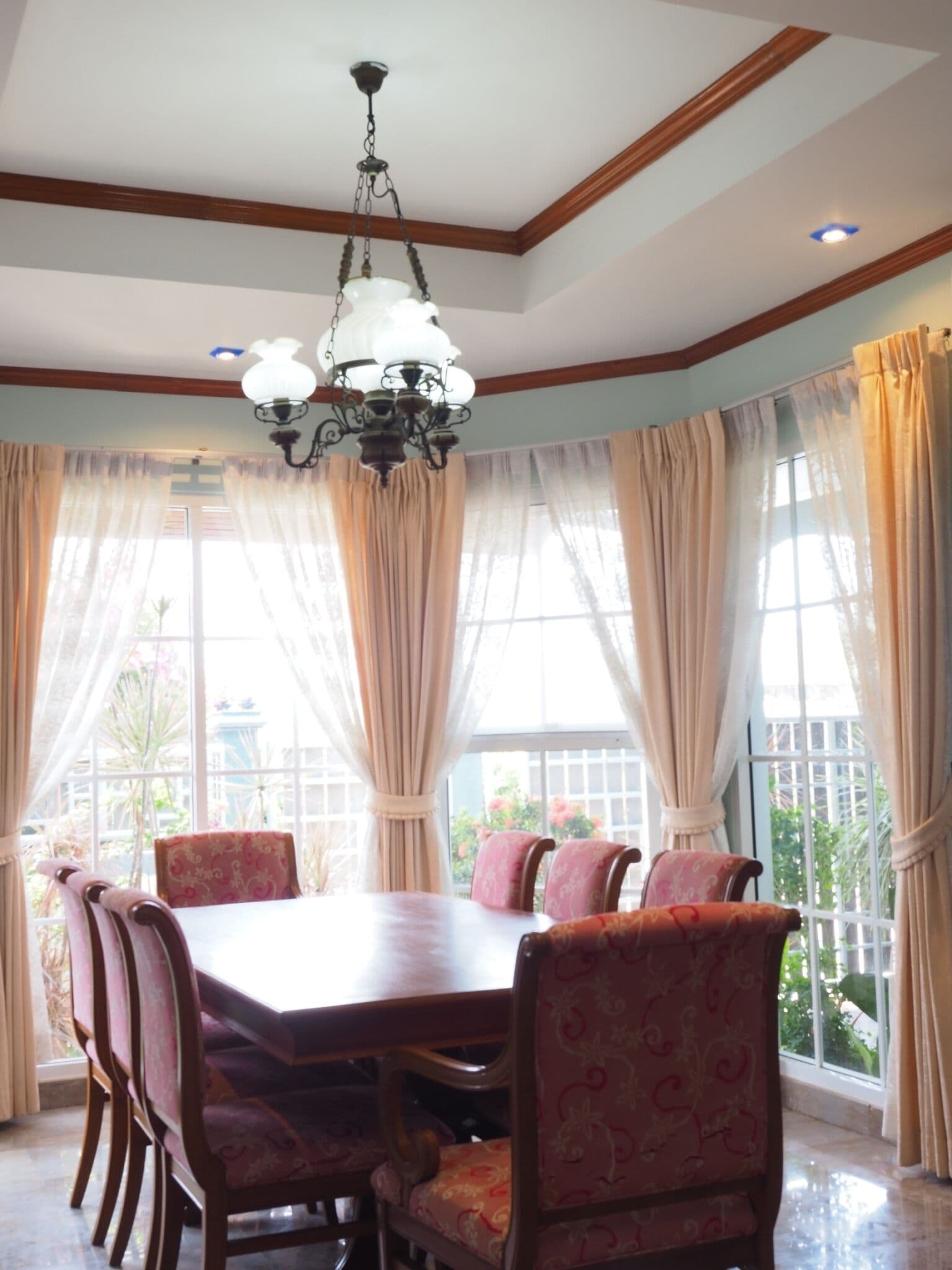 Un comedor con mesa y sillas de madera, ventanas amplias con cortinas y una lámpara de araña.
