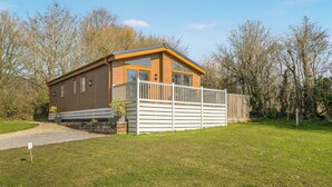 Une petite maison en bois d’un seul étage, dotée d’un balcon, entourée d’une cour verdoyante et d’arbres.