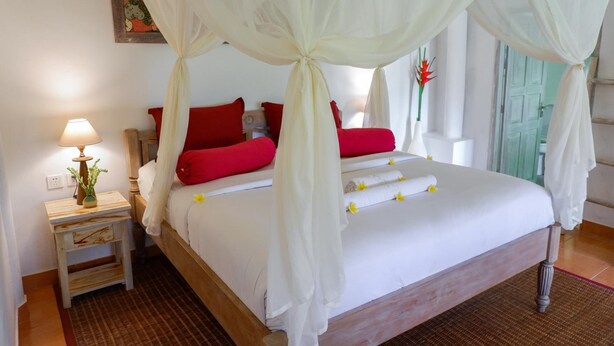 Una cama con dosel, almohadones rojos, una mesita de noche con lámpara y una pequeña planta.
