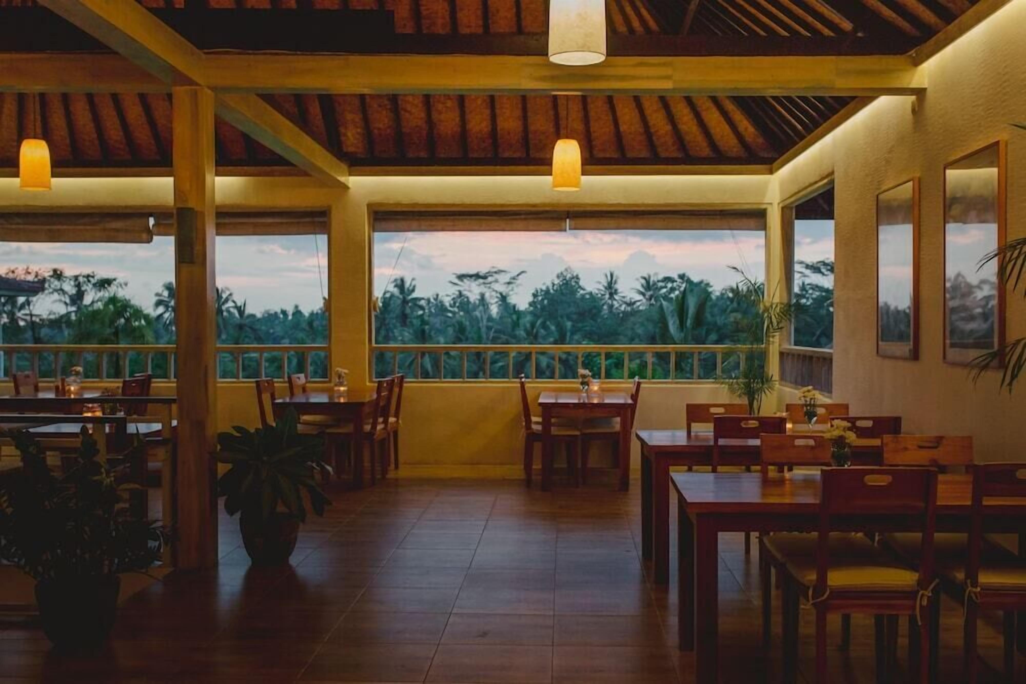 Um restaurante com mesas e cadeiras de madeira, grandes janelas com vista para palmeiras e luminárias pendentes.