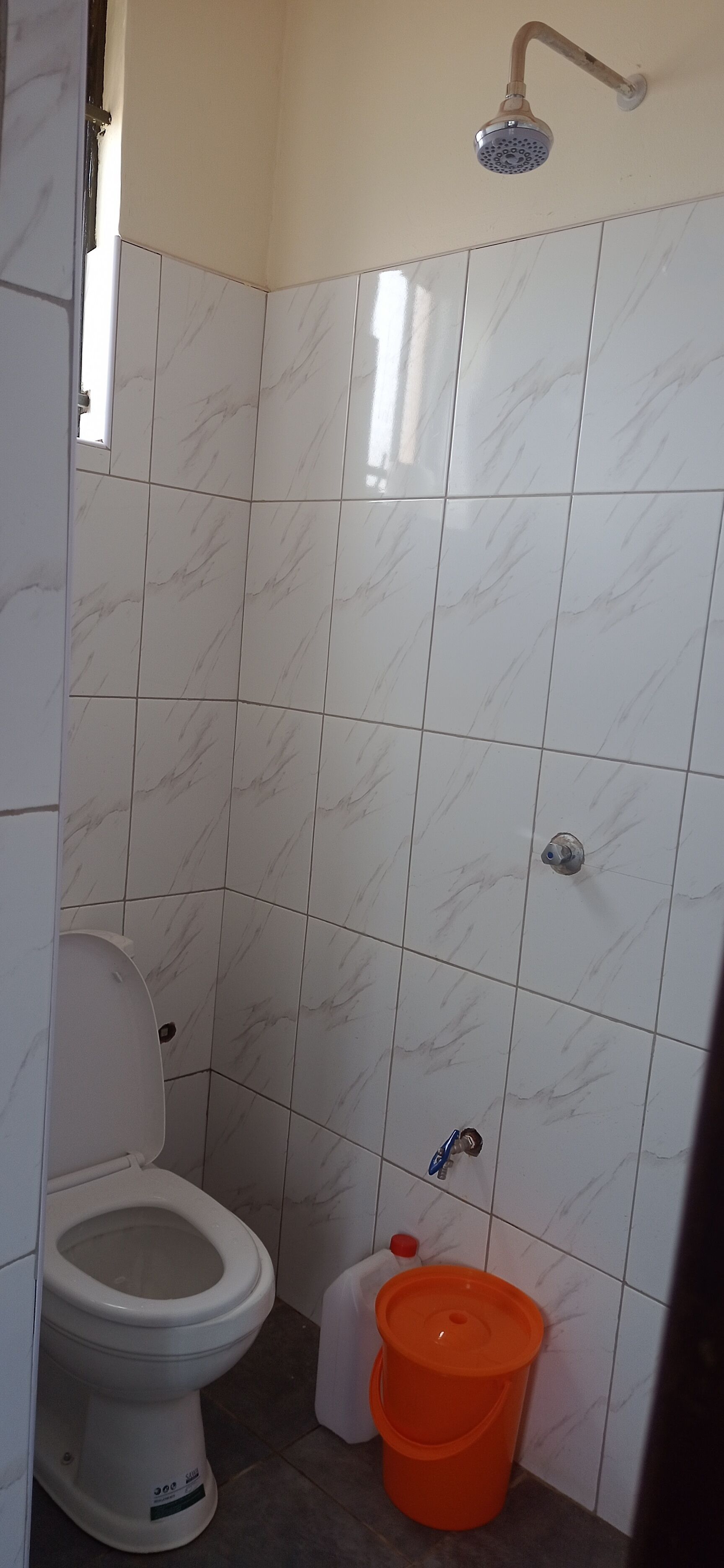 A bathroom with white marble tiles, a white toilet, a showerhead, and an orange bucket.