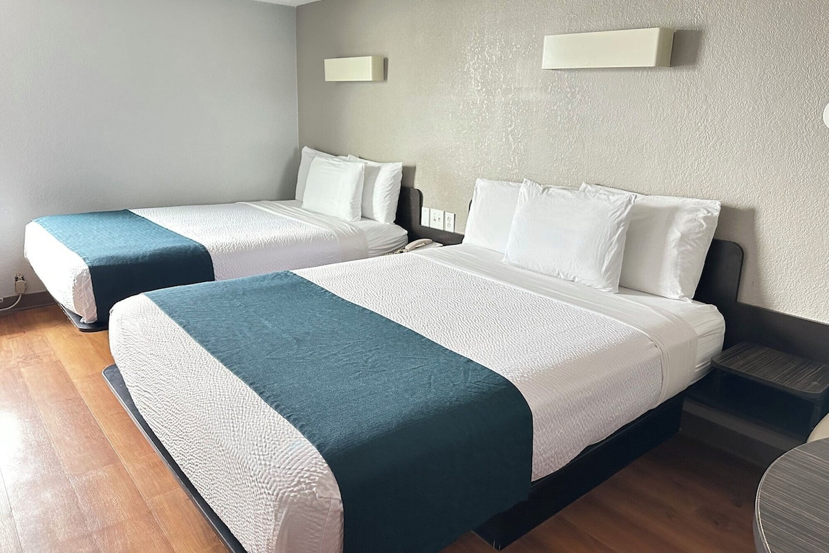 A hotel room with two beds, each with white bedding and teal accents. There is a small bedside table with a lamp, a wall-mounted light, and a power outlet.