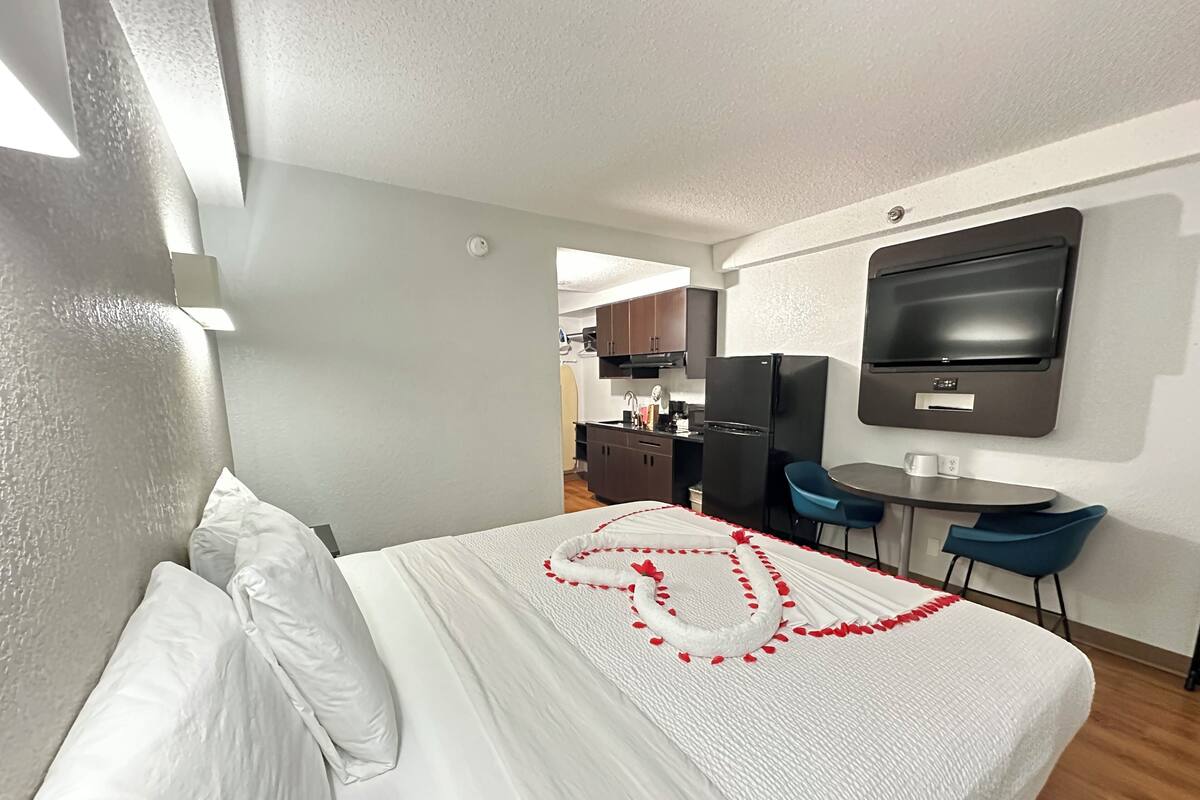 A modern hotel room with a neatly made bed, a small kitchenette, a flat-screen TV, and a dining area with blue chairs.