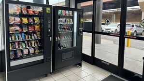 Máquina expendedora llena de aperitivos y bebidas, ubicada en un edificio con ventanas grandes y un estacionamiento visible.