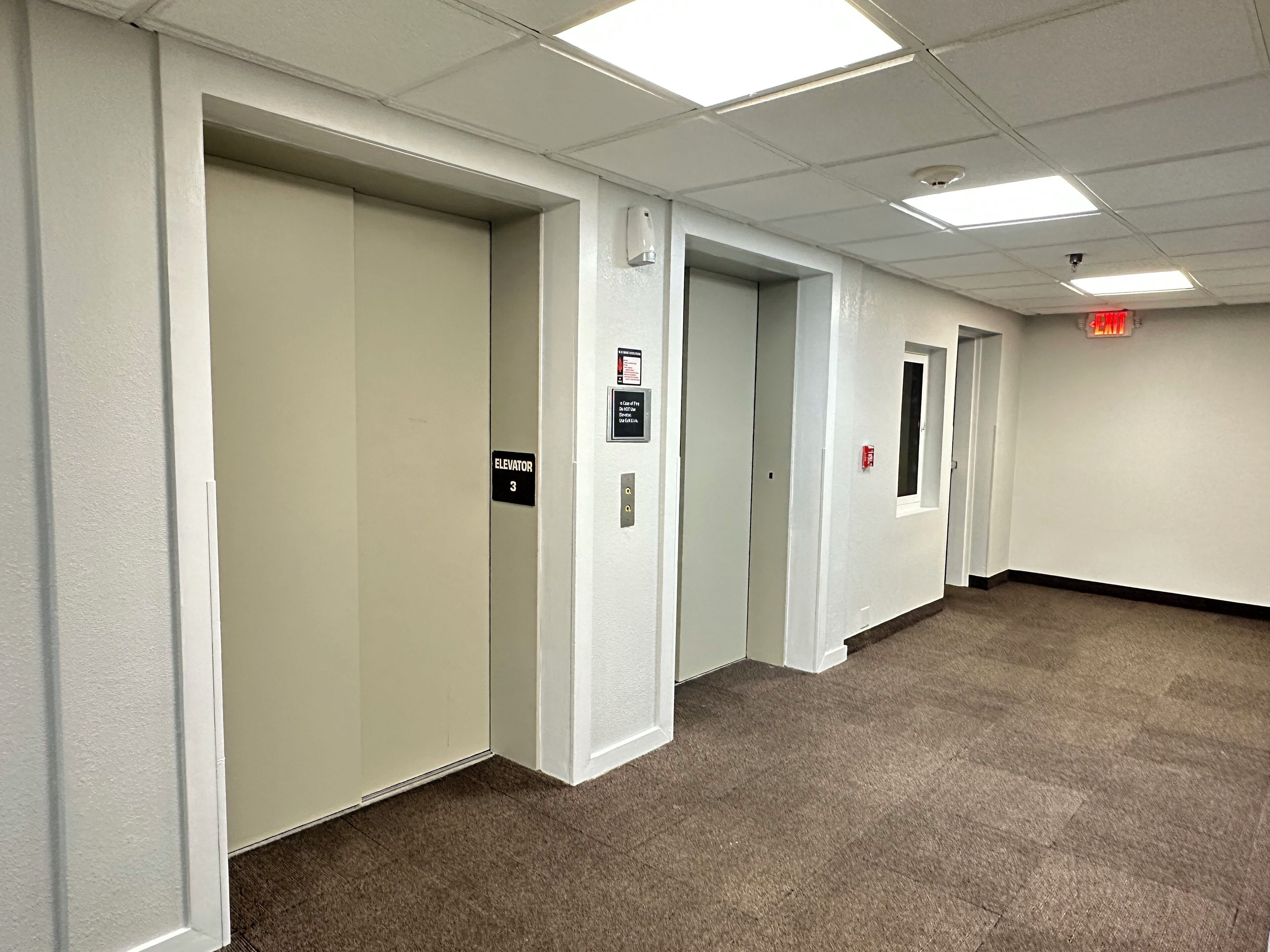 A hallway with three doors, each marked with an elevator sign, carpeted flooring, and fluorescent lighting.