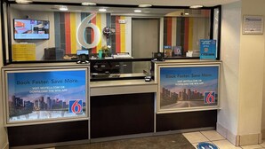 Un mostrador de recepción de hotel con un número '6' grande, una pantalla digital mostrando información de las habitaciones y carteles promocionales que animan a los huéspedes a reservar a través del sitio web o la aplicación del hotel.