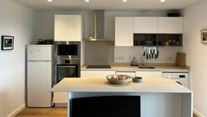 A modern kitchen with white cabinetry, a built-in oven, microwave, and dishwasher. There is a central island with a bowl of fruit and a kettle. - Renovated apartment with beautiful view of the beach golf course 5 minutes away (IB)