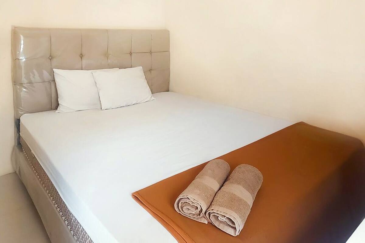 Cama bem arrumada com lençóis brancos e duas toalhas enroladas sobre um cobre-leito marrom.