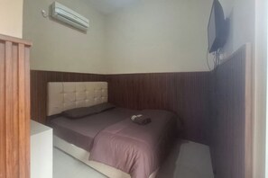 Una habitación pequeña con cama individual, cabecera de madera, televisor de pantalla plana montado en la pared y aire acondicionado sobre la cama.