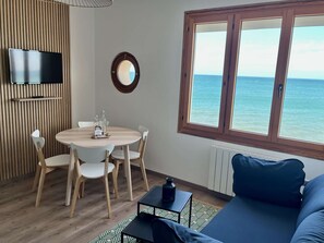 A room with a round wooden table, white chairs, a blue sofa, a TV mounted on a wooden panel, a window with a view of the sea, and a radiator.