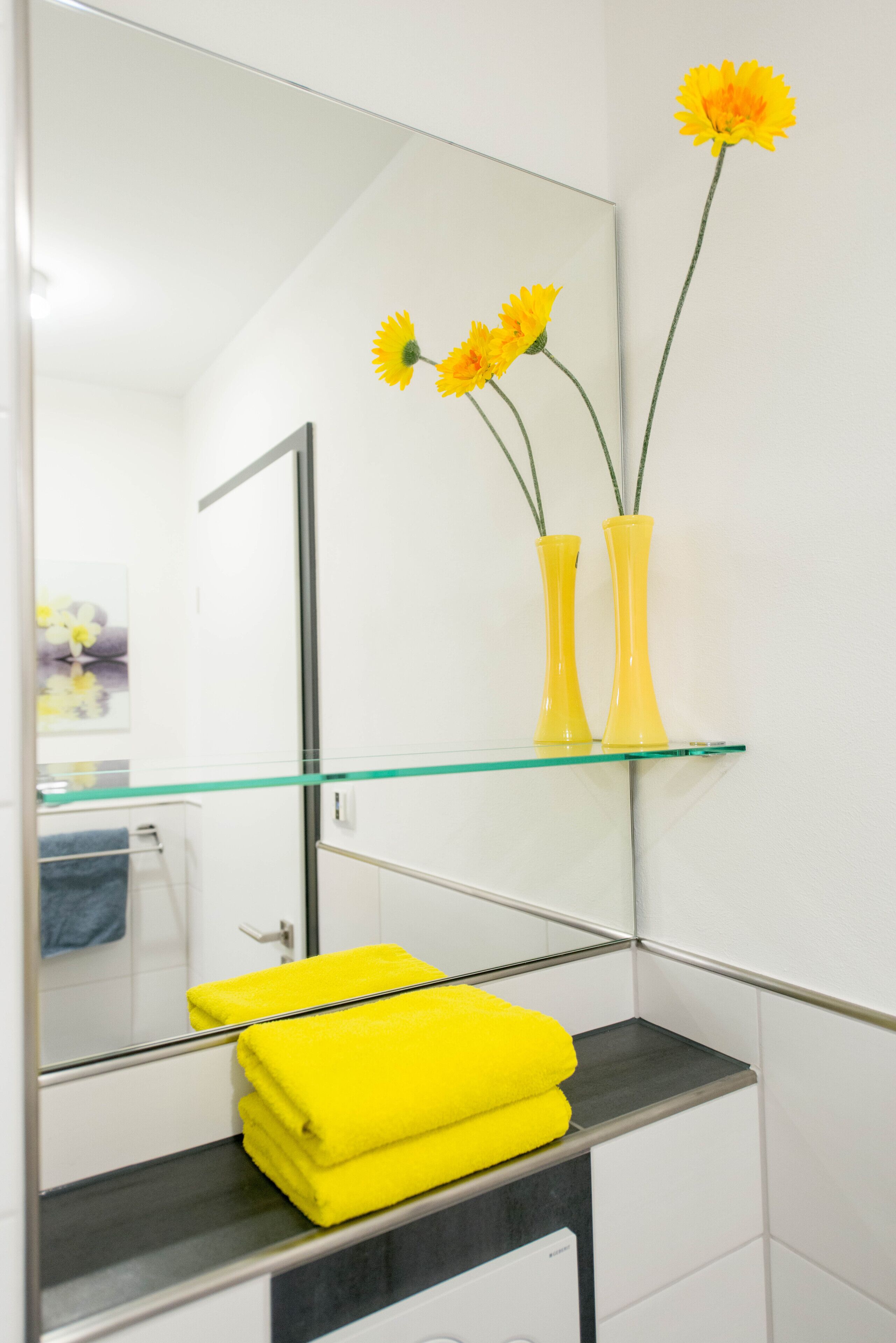 A bathroom with a glass shelf holding yellow towels and two yellow vases with flowers.