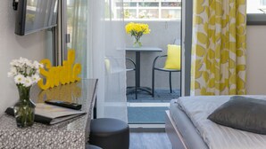 Una habitación de hotel con una cama, una mesita con un jarrón de flores, una silla y una ventana con cortinas amarillas.