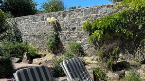 Two striped lounge chairs with cushions, a small table with a pitcher and glass, and a stone wall in the background.
