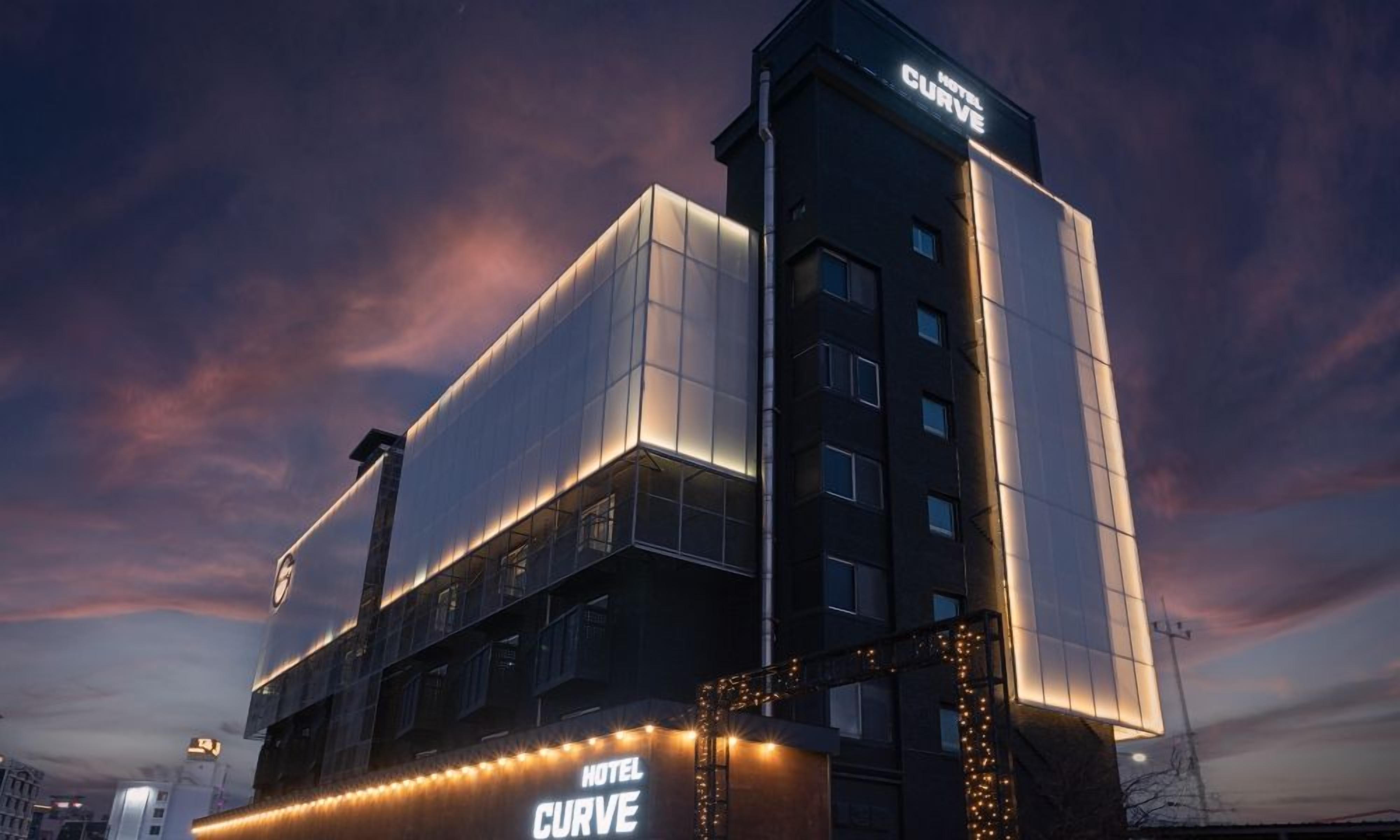 A modern hotel with a prominent 'HOTEL CURVE' sign, large windows, and a flat roof design.