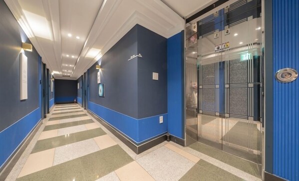 A hotel corridor with blue walls, a checkered floor, and a glass elevator. - CURVE HOTEL IKSAN (Iksan)