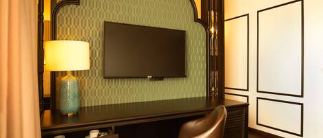 A hotel room with a dark wooden desk, a television mounted on a green patterned wall, a brown leather chair, and a lamp.