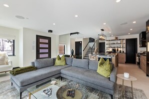 A modern living room with a grey sectional sofa, a glass coffee table, and a kitchen in the background.