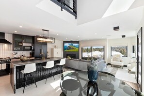 A modern kitchen with a central island, a dining area with a glass table, and a living space with a sofa and a large window.