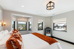 A modern bedroom with a large bed, multiple windows, a fireplace, and a view of the outdoors.