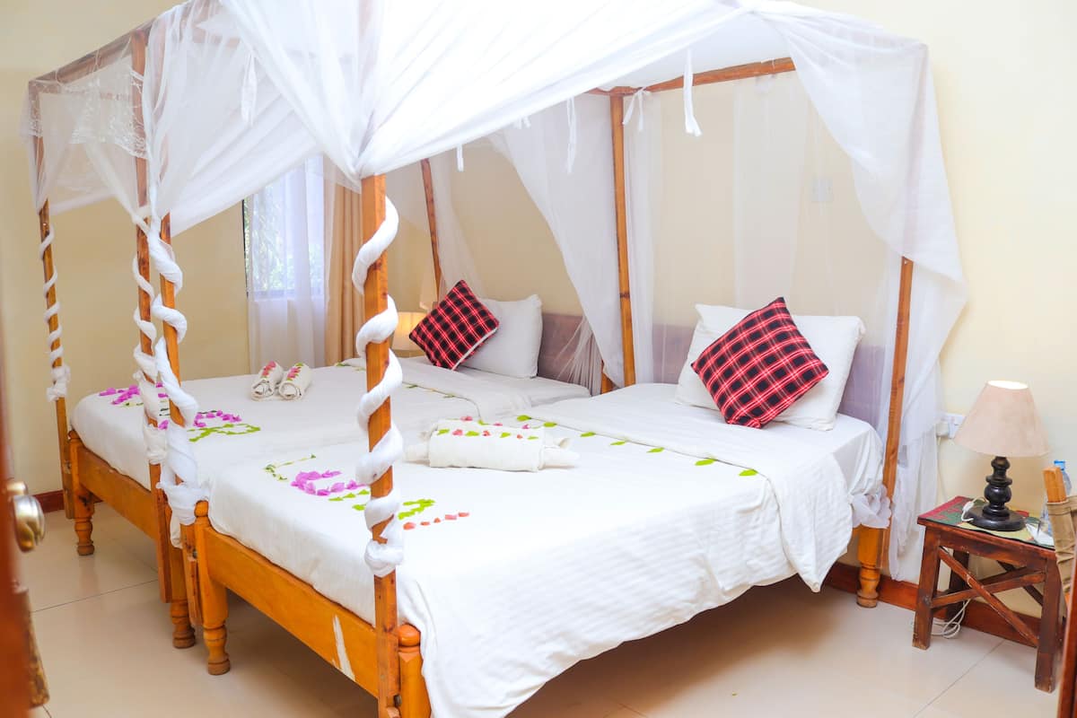Una cama con dosel y cortinas, ropa de cama blanca y adornos florales, una mesita de noche con lámpara y una pequeña mesita auxiliar de madera.