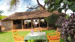 A thatched-roof hut with a stone wall, surrounded by a grassy area with wooden chairs and a table set for dining. - River Lumi Resort - Taveta (Chala)
